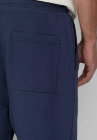 Navy blauwe joggingsbroek met een achterzak met gestikt kader en logo-accent. Zachte textuur, elastische tailleband, ontspannen pasvorm.