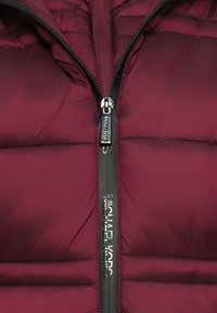 Maroon quilted puffer jacket with black waterproof zipper featuring silver pull tab and "MICHAEL KORS" branding along zipper line.