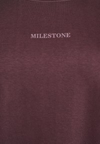 Burgundy cotton t-shirt featuring the word "MILESTONE" in light pink above the chest area. Smooth texture, crew neck design.