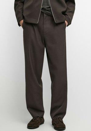 Persona che indossa pantaloni larghi marrone scuro, giacca scura abbinata e scarpe marroni con lacci, in piedi con le mani nelle tasche.