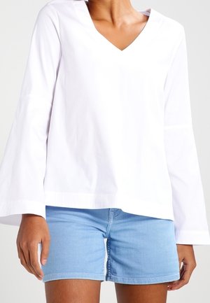 Person iført en hvid langærmet V-hals top med flagrende ærmer og lyseblå denimshorts, stående mod en ensfarvet baggrund.