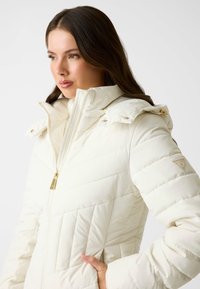 Giacca invernale bianca imbottita con colletto alto, chiusura con zip dorata e patch con il marchio sulla manica. Presenta un motivo a rombi diagonale e tasche.