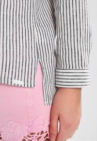 Camicia a righe bianche e nere con polsino ripiegato, fessura laterale e etichetta bianca. Abbinata a una gonna rosa con bordi in pizzo floreale.