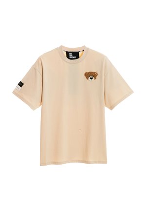 Persona con cabello rubio corto lleva una camiseta beige de gran tamaño con la cara de un oso de peluche grande en la espalda, pantalones cortos rojos, calentadores de piernas beige y zapatillas deportivas.