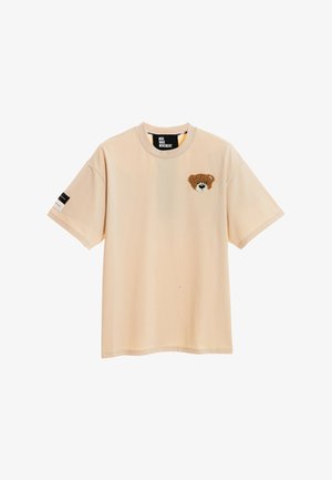 Persona con cabello rubio corto lleva una camiseta beige de gran tamaño con la cara de un oso de peluche grande en la espalda, pantalones cortos rojos, calentadores de piernas beige y zapatillas deportivas.