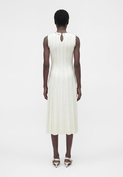 Γυναίκα με κοντά σγουρά μαλλιά που φοράει ένα λευκό πλεκτό μίντι φόρεμα χωρίς μανίκια με άνοιγμα στην πλάτη και μπεζ τακούνια, με φόντο μια μονόχρωμη επιφάνεια.