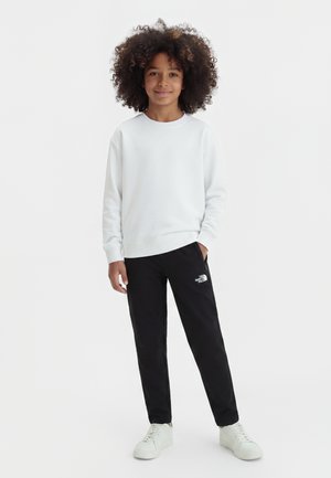 Enfant aux cheveux bouclés debout, portant une chemise blanche à manches longues, un pantalon noir North Face et des baskets blanches sur un fond uni.