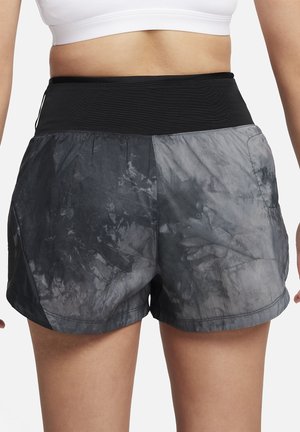 Osoba w białym staniku sportowym i czarno-szarych sportowych spodenkach typu tie-dye, pokazana od tyłu, stoi z rozluźnionymi ramionami.
