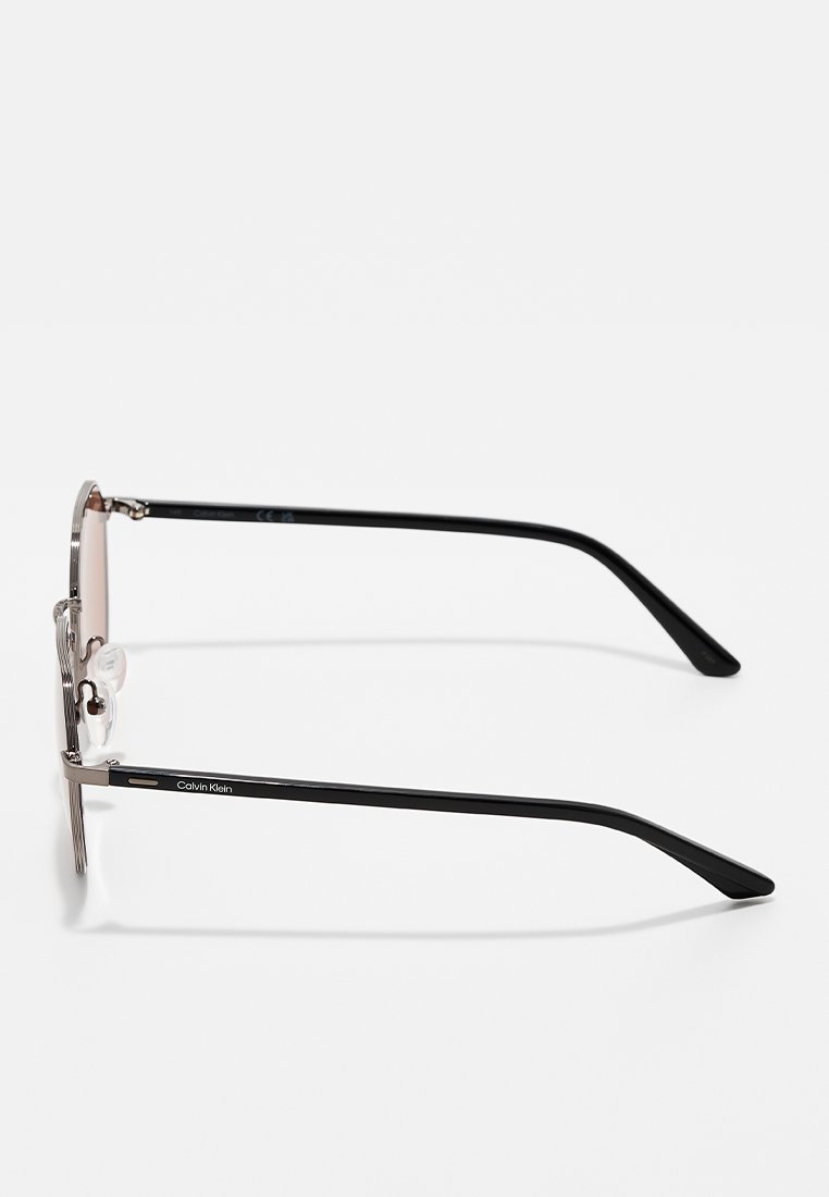 Occhiali da sole con montatura in metallo e lenti sfumate. Presentano aste nere e naselli trasparenti. Logo Calvin Klein visibile su un'asta.