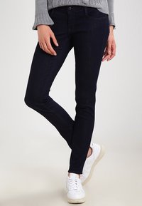 Mörkblå skinny jeans med en slät yta, femficksdesign och klassisk knapplåsning med dragkedja, i kombination med vita sneakers.