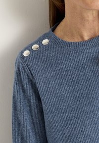 Frau trägt einen blauen gerippten Pullover mit drei silbernen runden Knöpfen an der Schulter in der Nähe des Halsausschnitts.