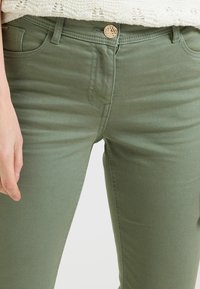 Jeans skinny vert olive en tissu mélangé coton, avec une fermeture à bouton doré, cinq poches et des détails de couture subtils.