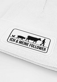 White knitted beanie featuring a rectangular black patch. The patch displays a graphic of a chicken, pig, cow, and a person, with text below.