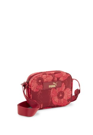 Czerwona torba typu crossbody Puma z dużym kwiatowym nadrukiem, regulowanym paskiem i złotą plakietką z logo z przodu na białym tle.