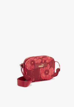 Czerwona torba typu crossbody Puma z dużym kwiatowym nadrukiem, regulowanym paskiem i złotą plakietką z logo z przodu na białym tle.