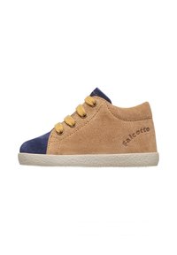 Chaussure suédée beige avec un bout en bleu marine, trois œillets dorés et une semelle en caoutchouc crème. Présente le nom de la marque embouti sur le côté.