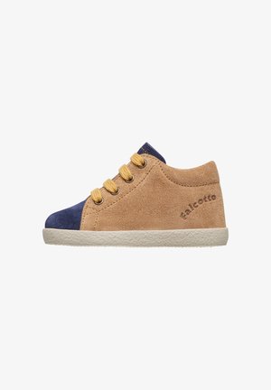 Chaussure suédée beige avec un bout en bleu marine, trois œillets dorés et une semelle en caoutchouc crème. Présente le nom de la marque embouti sur le côté.