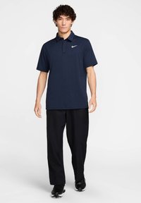 Tamsiai mėlynas polo marškinėlis su dviem mygtukais ant apykaklės, trumpomis rankovėmis ir baltu Nike logotipu. Suderinta su juodomis kelnėmis ir juodais sportiniais batais.