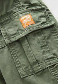 Tejido verde con superficie texturizada, que presenta una etiqueta de parche "SUPERDRY" en naranja. Incluye bolsillos y costuras cosidos.