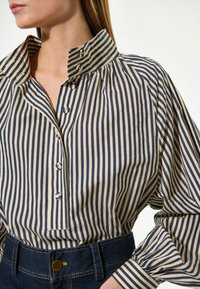 Gestreiftes Hemd in Beige und Marineblau, mit hohem Kragen, Knopfleiste und gerafften Ärmeln. Kombiniert mit einem dunklen Denim-Bund.
