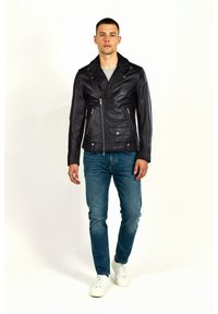 Mand iført en sort læderjakke, blå jeans, grå skjorte og hvide sneakers, der går på en ensfarvet hvid baggrund.