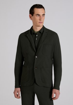 Man draagt een donker op maat gemaakt colbert en bijpassende broek, kijkt opzij tegen een effen grijze achtergrond.