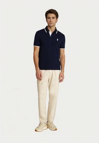 Young man stands confidently, wearing a navy polo with white trim and beige pants, styled with light-colored sneakers.