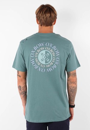 T-shirt en coton vert avec un design de logo circulaire en bleu clair et violet au dos. Manches courtes, col rond, coupe décontractée.