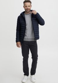 Jachetă puffer albastru închis cu glugă, pulover gri cu întreținere, pantaloni negri și ghete albe. Jacheta are fermoar în față și cusături orizontale.