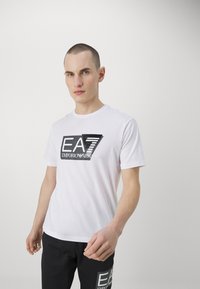 White cotton t-shirt featuring a black EA7 logo on the chest, short sleeves, and a crew neck design. Smooth texture, standard fit.