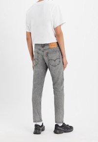 Jeans de mezclilla gris con un corte tapered, que presentan un parche de cuero de la marca y dos bolsillos traseros, combinados con una camiseta blanca corta y zapatillas negras.