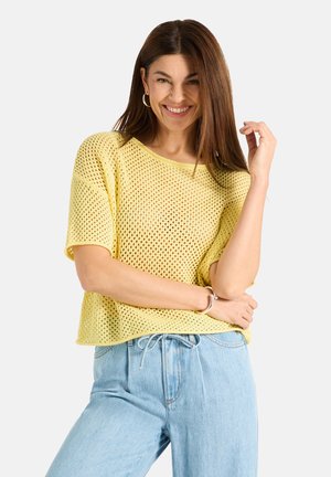 Lächelnde Frau mit langen braunen Haaren, die Creolen, ein gelbes kurzärmeliges Stricktop, hellblaue Jeans und ein silbernes Armband trägt, entspannt stehend.