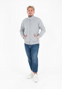 Giacca zip-up grigia chiara con colletto alto, dotata di tasche laterali. Indossata con jeans in denim blu e sneakers bianche. Design semplice e casual.