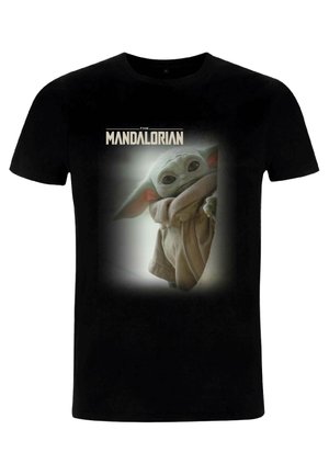 T-shirt noir ornée d'un graphique représentant un personnage avec de grandes oreilles et une peau verte, vêtu d'une robe beige, encadré par un doux effet de halo.