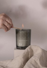 Scented candle in a translucent gray glass container with a geometric pattern, lit with a wooden stick, beside a folded linen cloth.