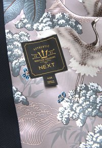 Hedvábná kravata s jemným fialovým pozadím a modrými květinovými a jeřábími vzory. Obsahuje černou nálepku s textem "Authentic Heritage Tailoring".
