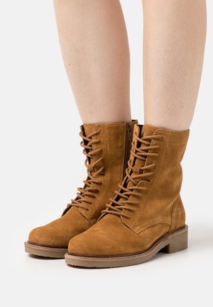 Botas de tornozelo em camurça castanha com um fecho de atacadores na frente e detalhe de zipper. Bico quadrado e sola de borracha ligeiramente elevada. Superfície texturizada.