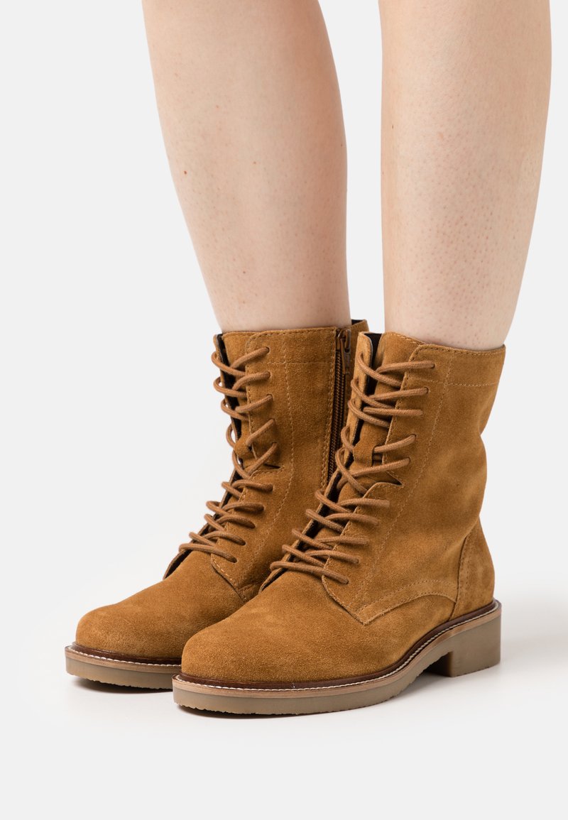 Botas de tornozelo em camurça castanha com um fecho de atacadores na frente e detalhe de zipper. Bico quadrado e sola de borracha ligeiramente elevada. Superfície texturizada.