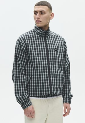 Tânăr purtând o jachetă cu fermoar în carouri alb-negru, cu guler înalt și pantaloni deschiși la culoare, uitându-se în lateral.