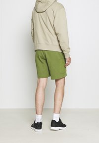 Beige mikina s kapucí, zelené kraťasy, boční kapsa a černé tenisky s bílou podrážkou a ponožkami. Jednoduché pozadí.