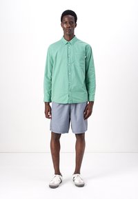 Mintgrünes Hemd mit Knöpfen, lange Ärmel, Brusttasche; kombiniert mit hellgrauen Shorts und weißen Turnschuhen. Einheitlicher Hintergrund.