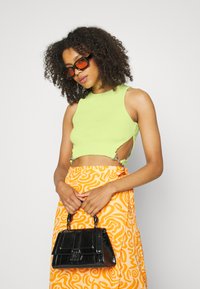 Top cropped verde limão com recortes laterais, saia com padrão laranja e uma bolsa preta em verniz com formato estruturado e alças curtas.