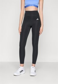 Sorte højtaljede leggings lavet af strækbar stof med en glat tekstur, med et lille hvidt logo på venstre lår, parret med hvide sneakers.