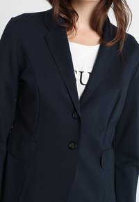 Marinblå blazer med en figursydd design, enfaldiga knappar, slagkrage och två framfickor. Slät textur och skräddarsydd form.