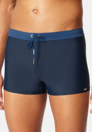 Męski tors w granatowych kąpielówkach ze ściągaczem zawiązanym na kokardkę w talii.