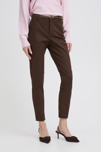 Sieviete valkā pieguļošas brūnas bikses, atbilstošu jostu, tumši brūnas kurpes ar smailiem purngaliem un gaiši rozā garām piedurknēm džemperi.