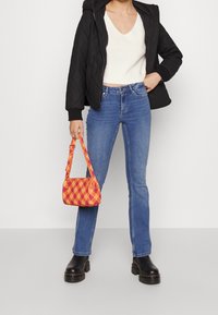 Veste noire matelassée, haut en maille blanc, jean évasé bleu et petit sac à main à carreaux orange et jaune. Bottes noires à semelle épaisse.