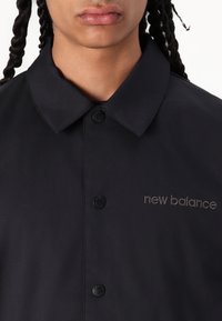 Camisa negra con cuello puntiagudo, que presenta botones a presión negros y el logotipo bordado de "new balance" en gris en el lado izquierdo. Textura suave.