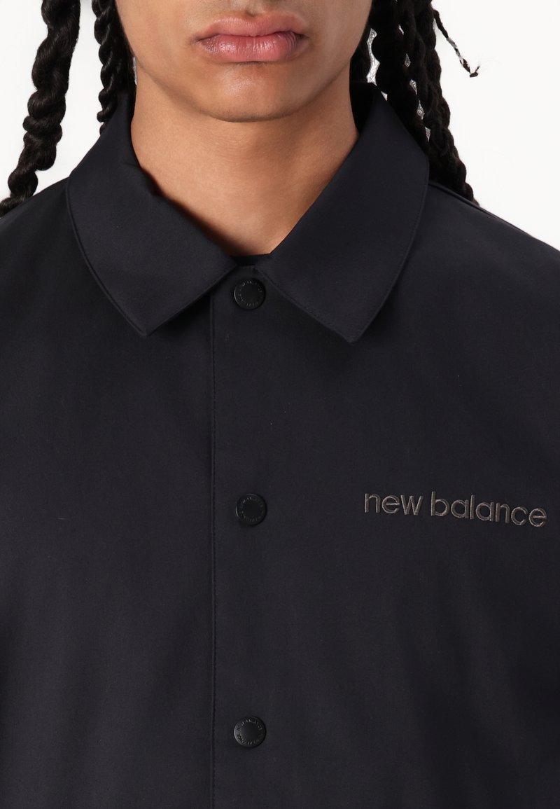 Camisa negra con cuello puntiagudo, que presenta botones a presión negros y el logotipo bordado de "new balance" en gris en el lado izquierdo. Textura suave.