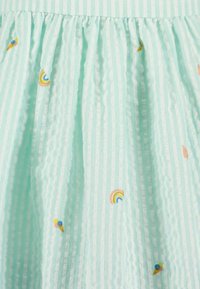 Le tissu de robe vert menthe présente des rayures verticales, des plis texturés et des détails brodés tels que de petits arcs-en-ciel et des cornets de glace.